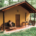 A girl is sitting on the front porch of a yellow house, which is located in the woods, and is taking a photograph.