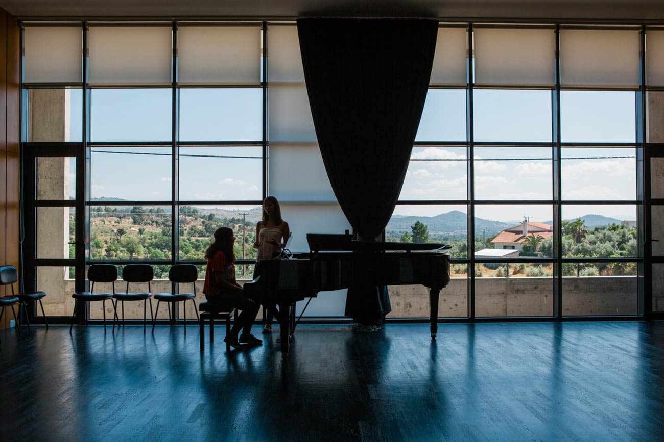Full color slideshow photo of Bendada Music Festival founder Ines Andrade leading a piano lesson with a student in the Casa Da Musica Da Bendada concert hall. The student, sitting at a baby grand piano, and teacher are silhouetted against floor to ceiling windows with a vista view of Bendada and the distant Portugal mountains.