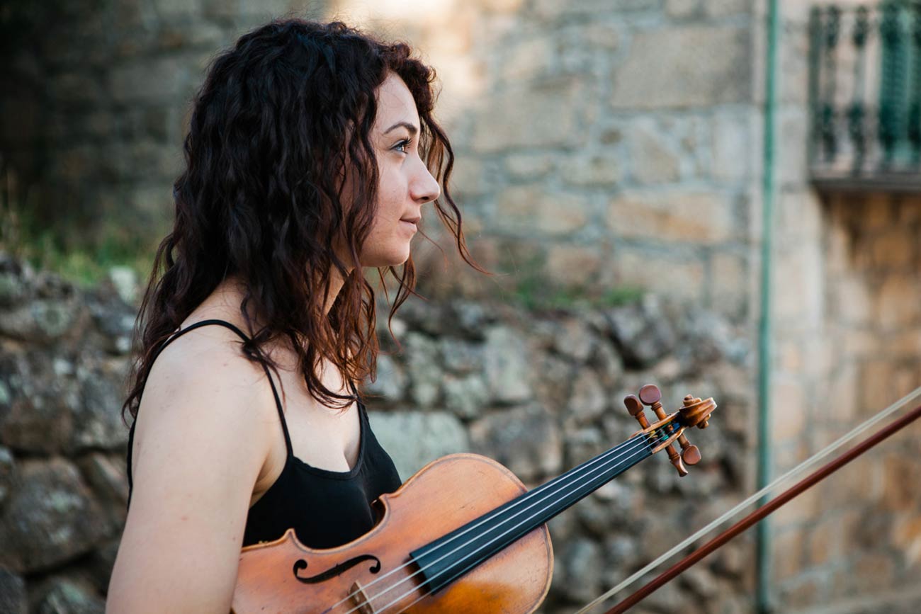 Full color slideshow profile portrait of Francesca Baldo, a violin student from Padua, Italy, holding her violin and bow as she watches her fellow music students perform in Bendada's village square.
