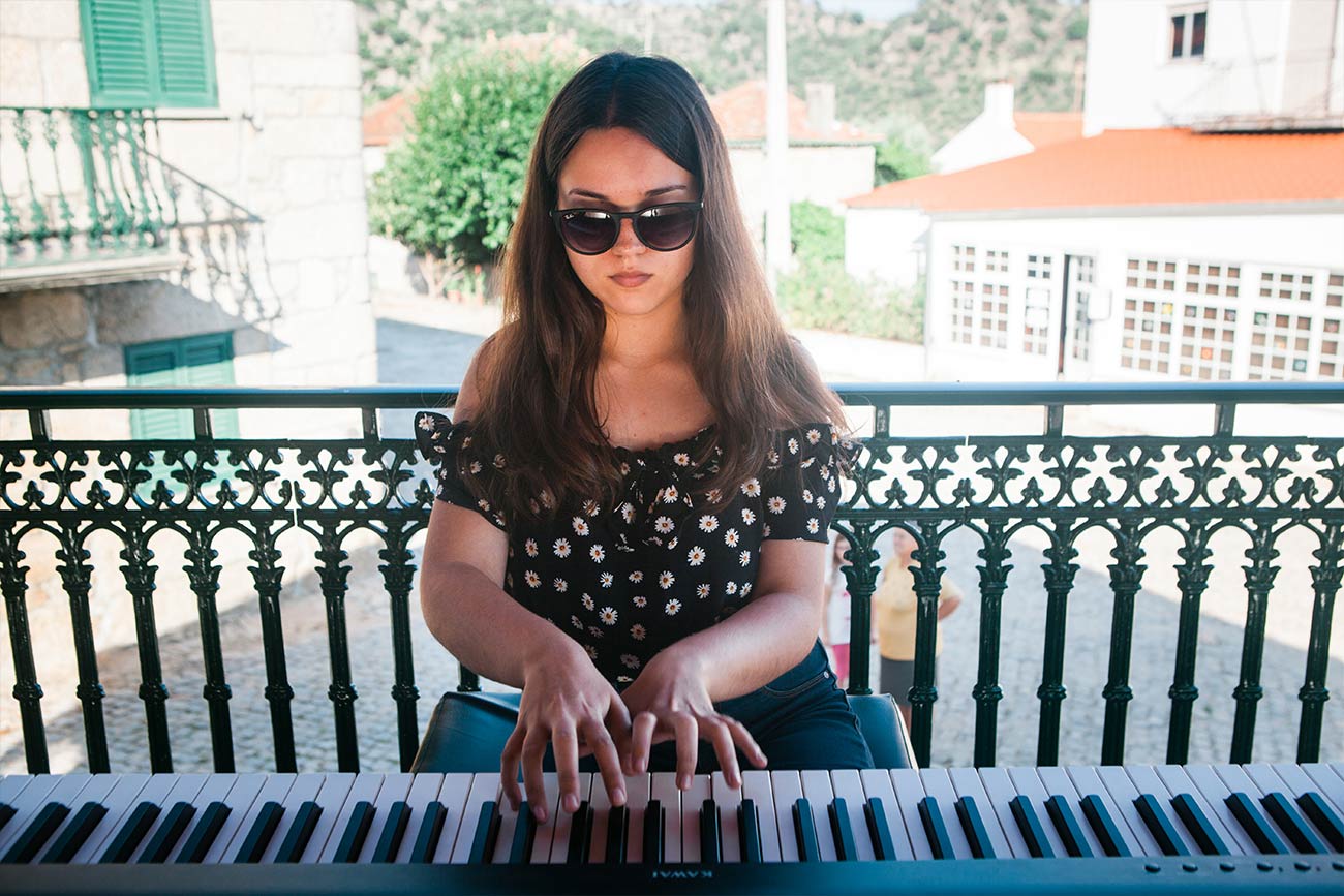Full color slideshow photo of piano student Margarida Pacheco wearing sunglasses, performing on a digital piano for the villagers of Bendada in the coreto, a covered bandstand in the town square.