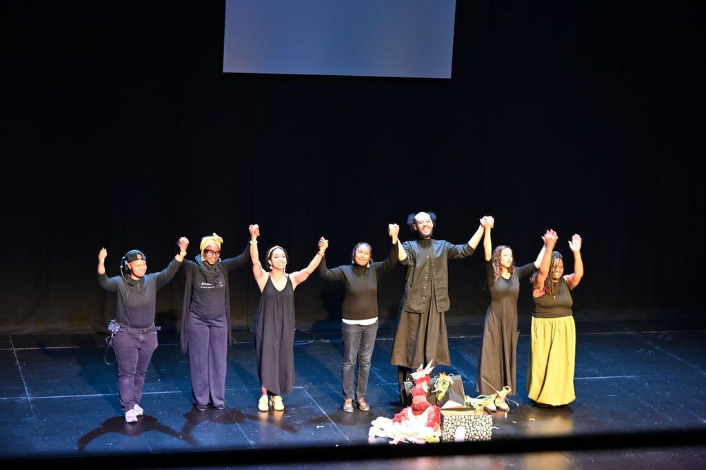 A group of seven people standing on a stage holding hands and raising them in a gesture of celebration or acknowledgment. They are dressed in mostly dark clothing, except for one person wearing a yellow skirt. In front of them on the stage are colorful props and items, including a red and white stuffed animal resembling a llama or alpaca. The background is dark with a large screen or panel partially visible above them.