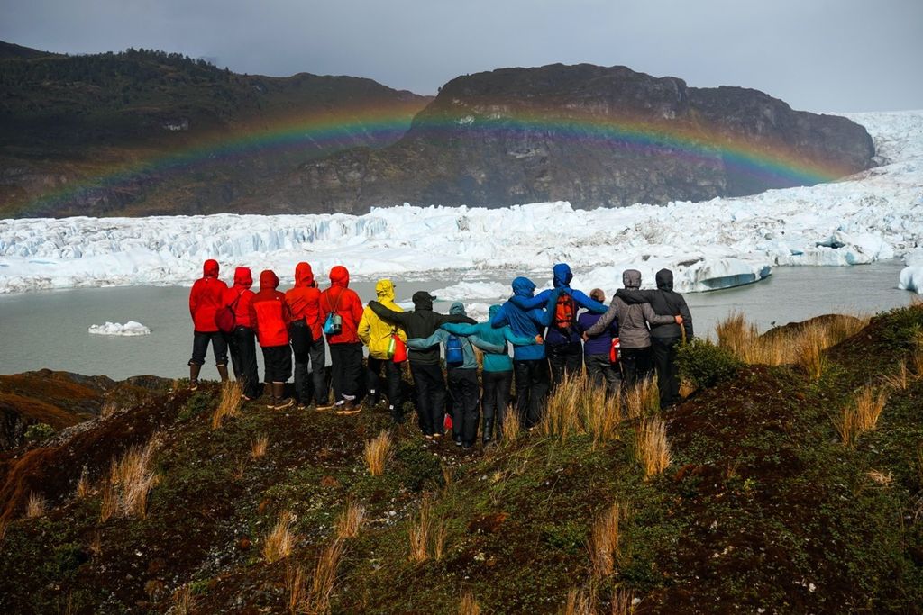 A group of people, bundled up in warm coats and backpacks, look out an ice bank with a rainbow above it