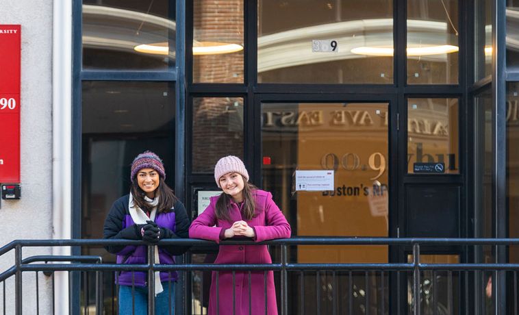 Two young women posing outside in winter hats and coats while leaning against a railing.