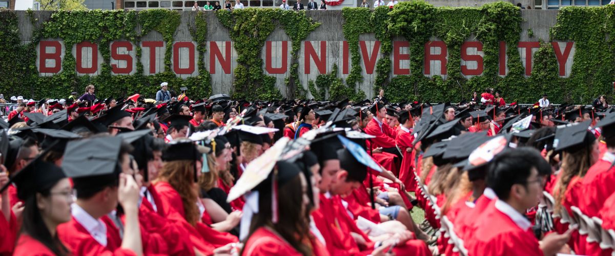 Side view of seated graduates in regalia by the Boston University sign.