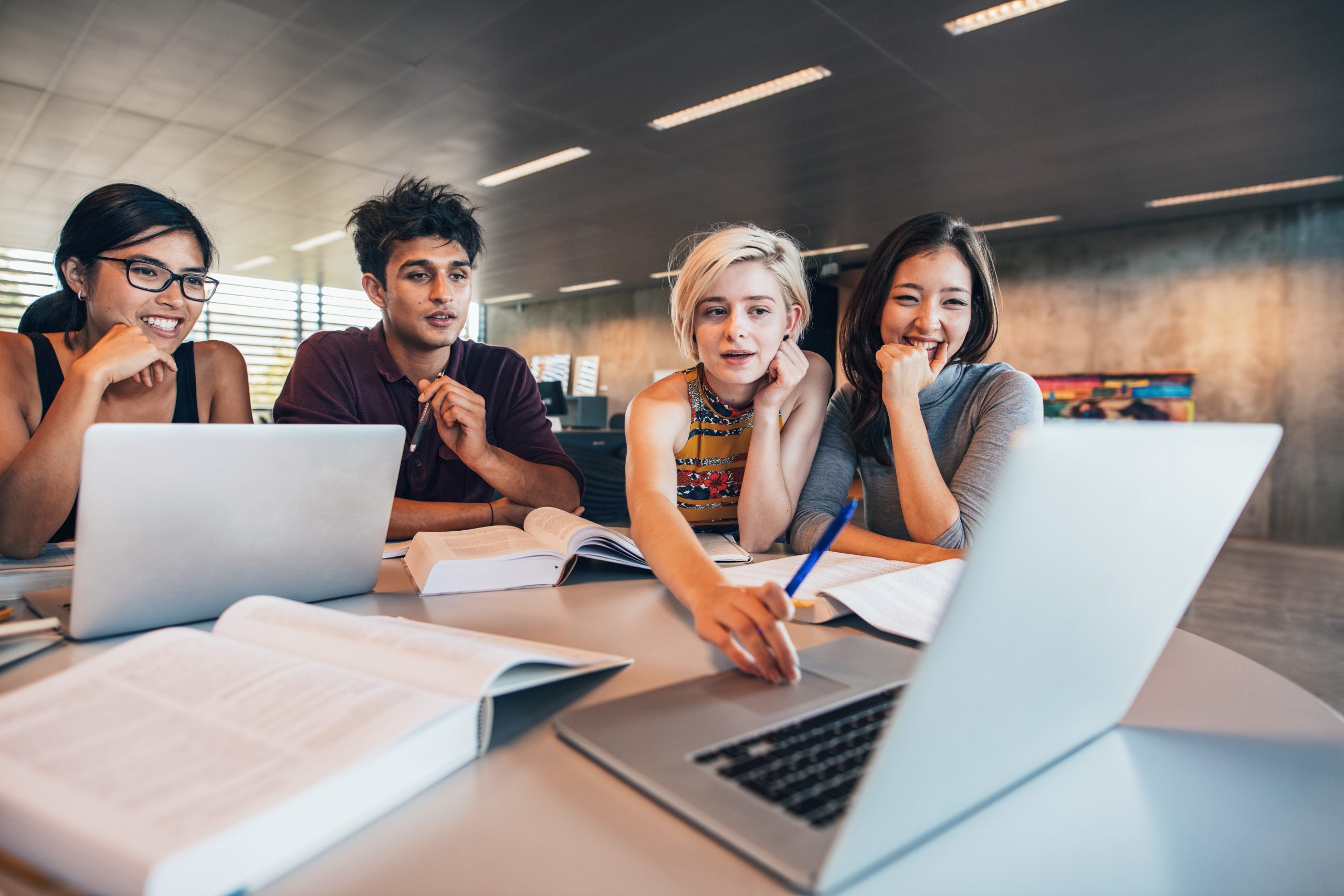 View from across: Two male and two female students working and collaborating in front of laptop. One female students reaching over and moving the cursor on laptop. Students are smiling.