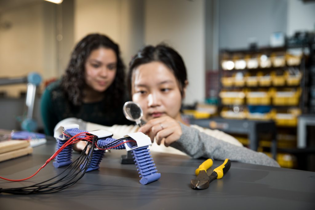 Student examining robotics work with a magnifying glass while a second student looks over her shoulder
