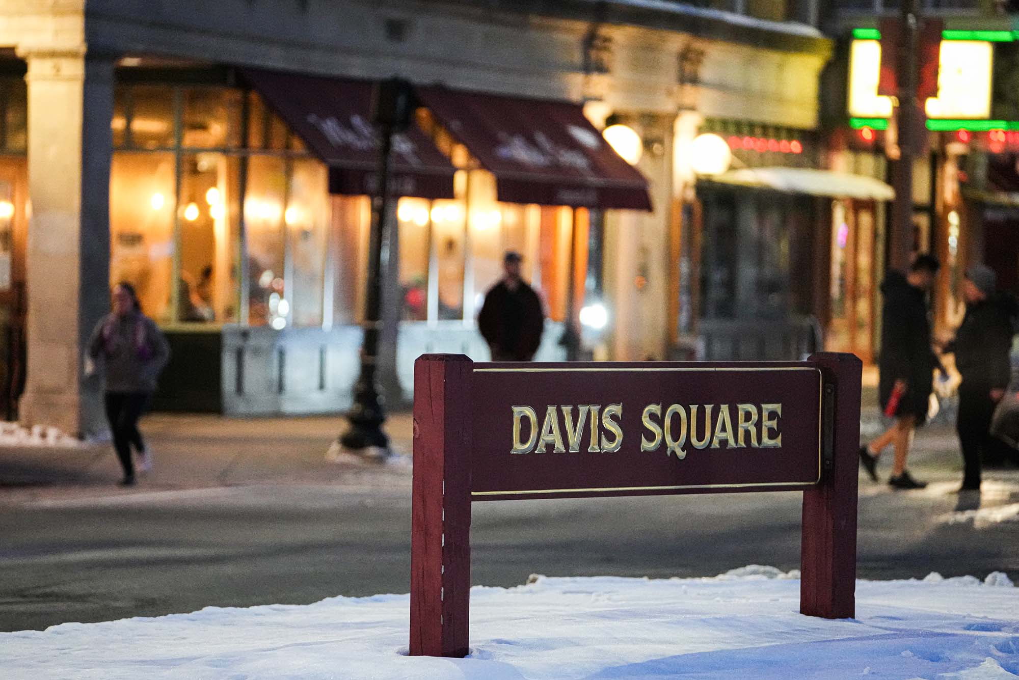 Photo of the red Davis Square sign with gold lettering taken when there was snow on the ground. Behind the sign, businesses are lit up at night.