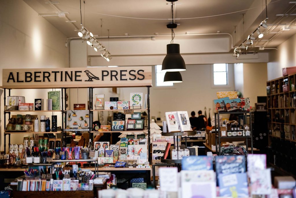 Photo of the front of Albertine press, which features wooden shelves and tables of trinkets, primarily paper goods and writing utensils. A wooden sign with black writing reads "Albertine Press"