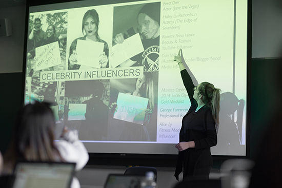 Elle Hvozdovic (COM’17)  stands in front of a projector screen