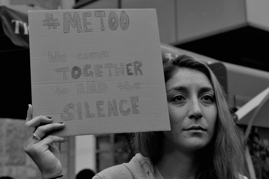 A woman attending a #MeToo Survivors March & Rally holds a cardboard sign reading, "We come together to end the silence."