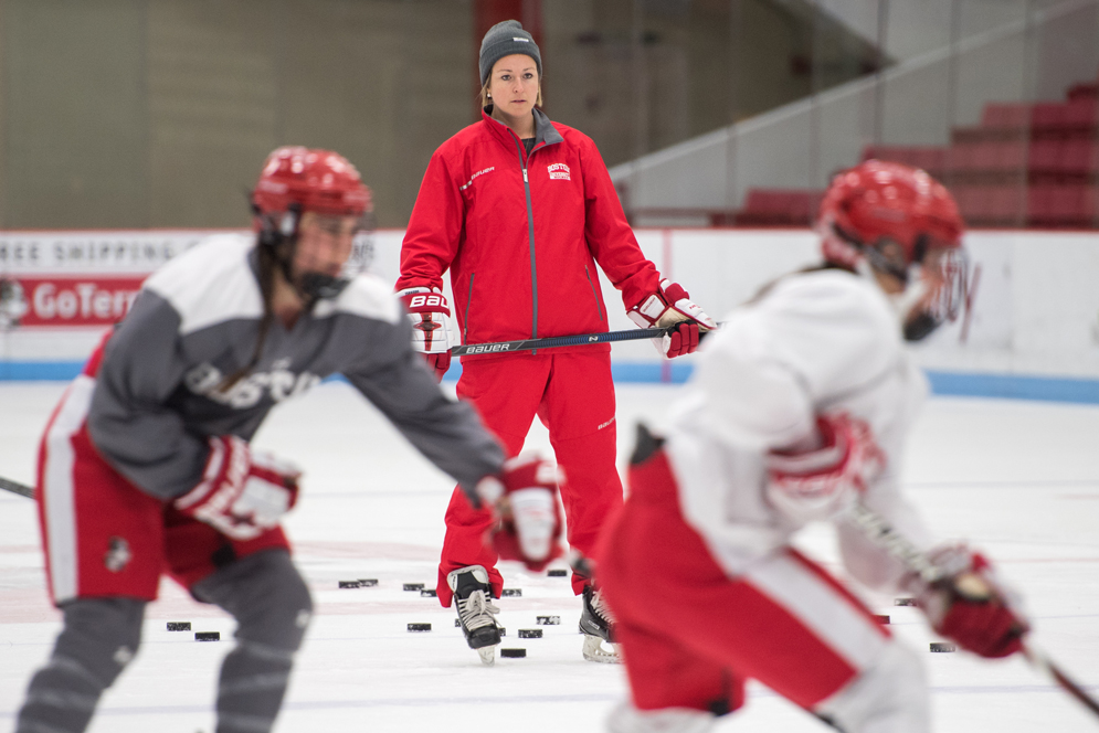 Coach Tara Watchorn stands on the ice with a hockey stick while she watches her players practice. 