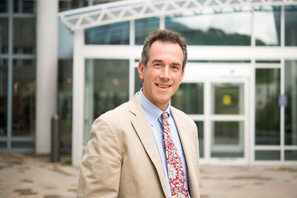 Portrait of Andrew Budson in a tan suit jacket and floral tie in front of a building with a white facade.