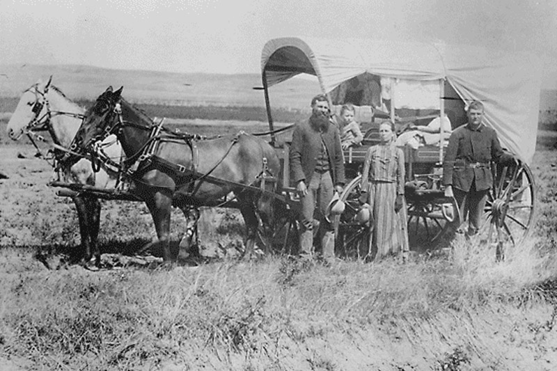 A family during the Great Western Migration, 1866, stands alongside their covered wagon.
