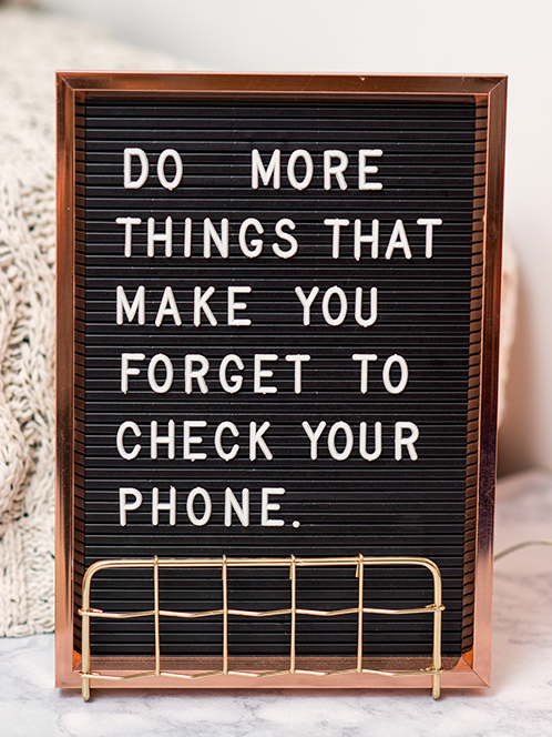 A black felt message board standing on a desk displays the message, 'DO MORE THINGS THAT MAKE YOU FORGET TO CHECK YOUR PHONE'