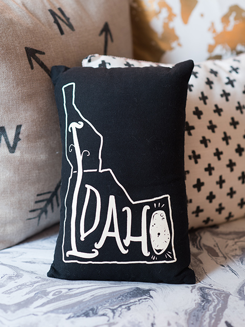 A black pillow painted with the shape of the state of Idaho and the word 'IDAHO' inside it sits on a multi-colored couch.