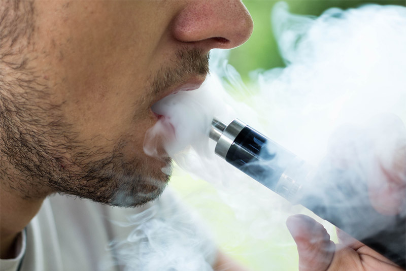 Tightly cropped photo of a young man vaping with an e-cigarette and vapor streaming out his mouth.