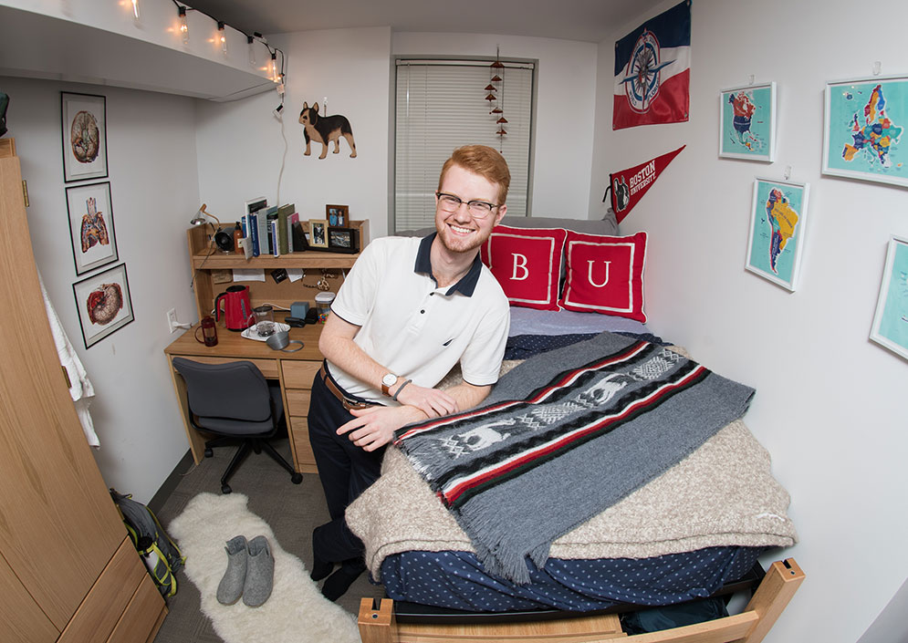 BU student Evan Dann stands smiling in his dorm room.