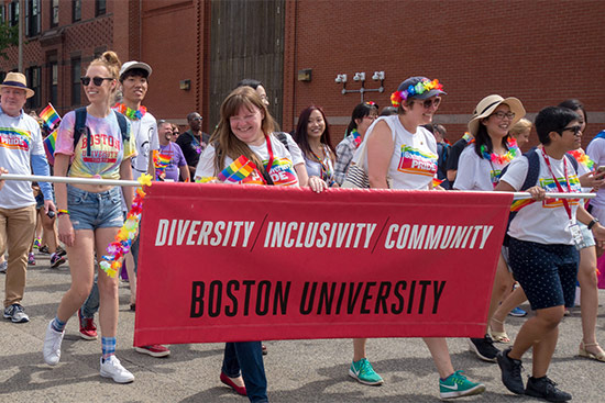 Boston University faculty and staff members march in the Boston Pride parade wearing t-shirts with the slogan 'Boston University Pride' and holding a banner with the slogan 'Diversity Inclusivity Community, Boston University'.