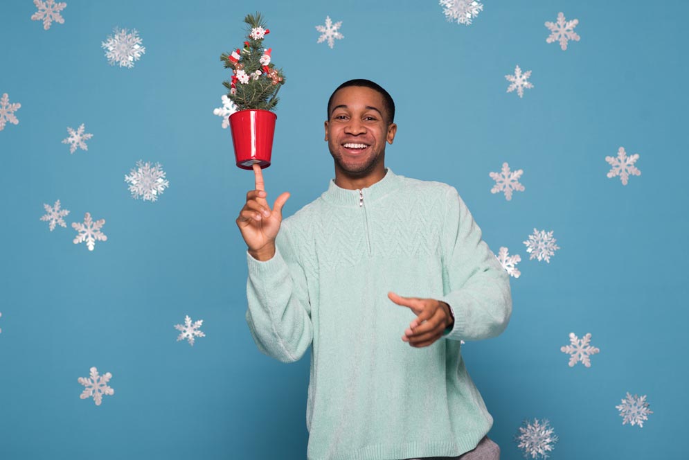 Kamali Chambers wears an ugly light blue holiday sweater while balancing a small christmas tree in his hand