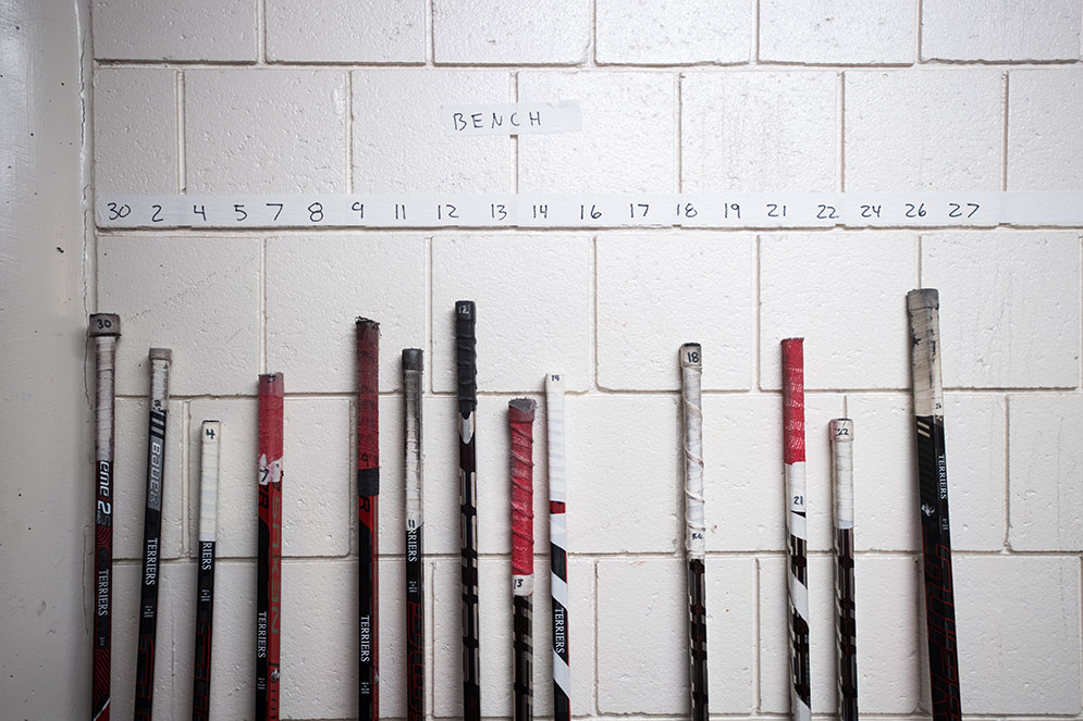Hockey sticks for the BU women's ice hockey players lean against the locker room wall, each beneath the number of the player it the stick belongs to.
