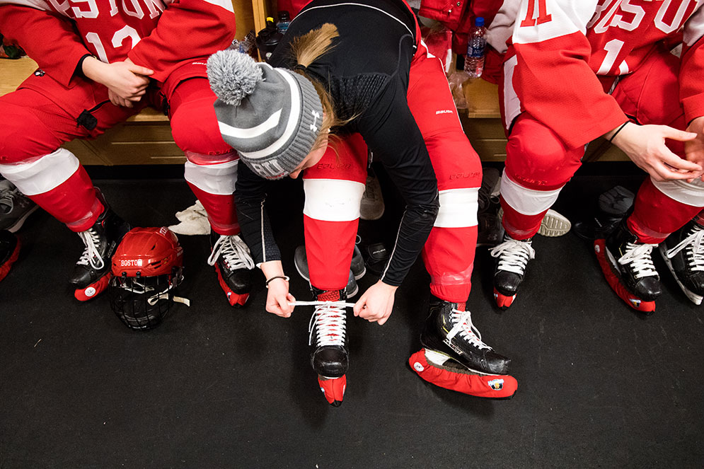A BU women's ice hockey player laces up her skates in the locker room.