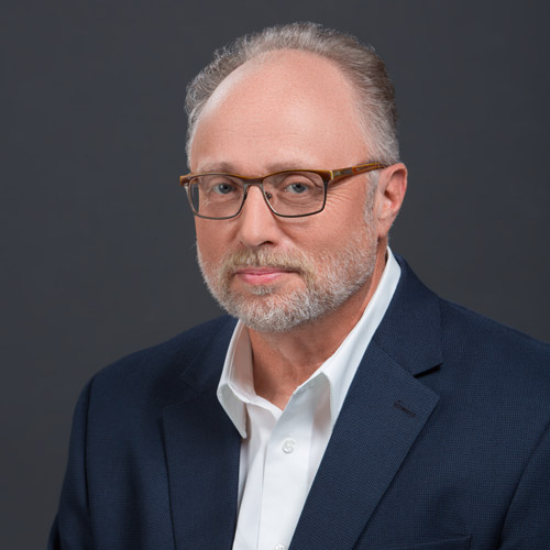 Portrait of Joel Brown. An older white man with greying brown hair, beard, and mustache and wearing glasses, white collared shirt, and navy blue blazer, smiles and poses in front of a dark grey background.