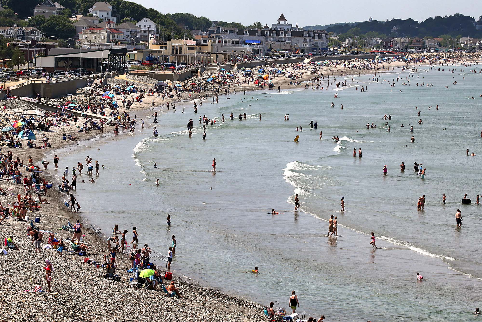 Wide photo of visitors crowding onto Nantasket Beach in Hull, MA. People are seen sitting on the small, partially rocky shore at left, while others are in the shallow water at right. Houses and buildings lining the street along the beach are seen in the background.