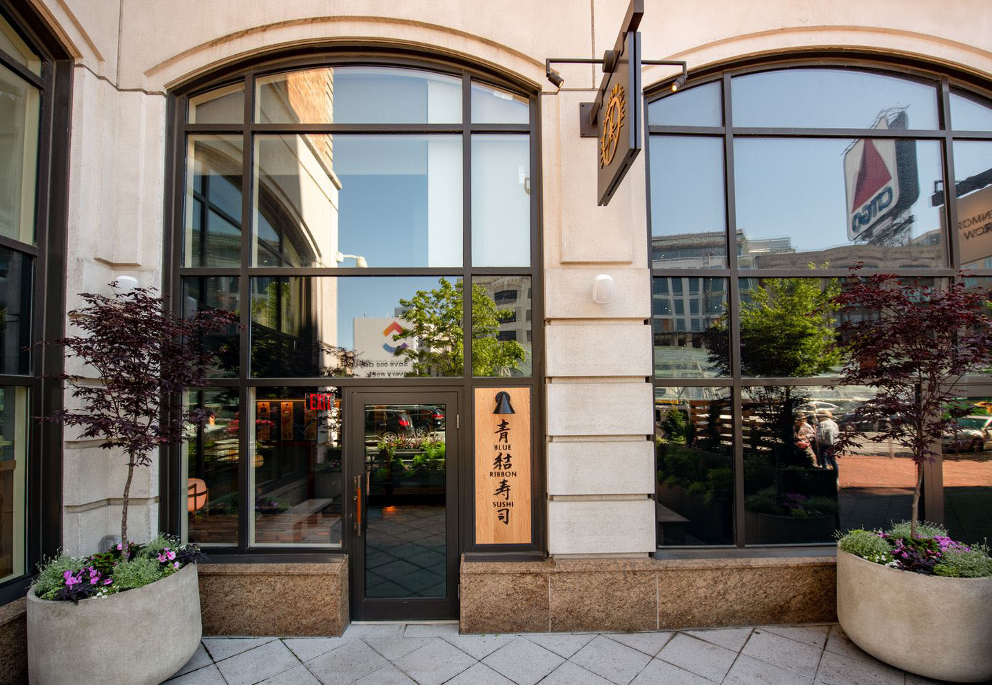 Photo of the exterior of Blue Ribbon Sushi restaurant. Large bay windows hosts a door with a wooden sign for the restaurant entrance.