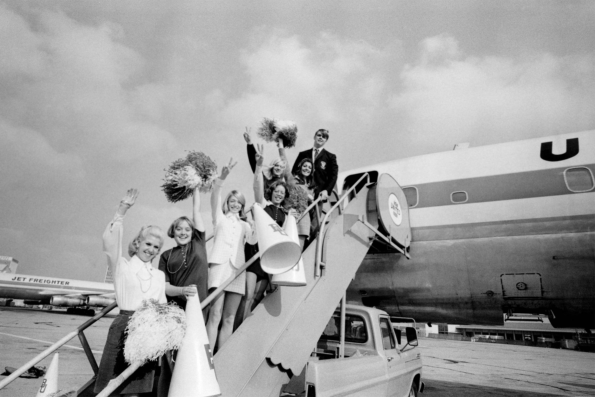 1969 Boston University football team captain Pat Hughes poses for a photo with BU cheerleaders while boarding a flight to California for the Pasadena Bowl game.