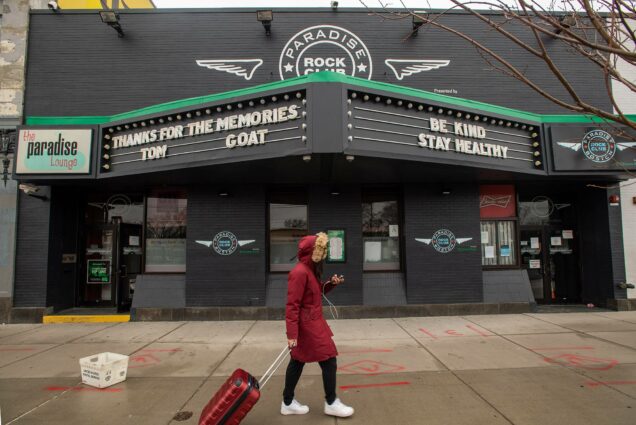 Young person with suitcase walks on the sidewalk. The Paradise Lounge's marquee displays the words "Thanks for the memories, Tom GOAT," and "Be Kind. Stay Healthy". The lounge will be closed for the duration of the coronavirus situation.