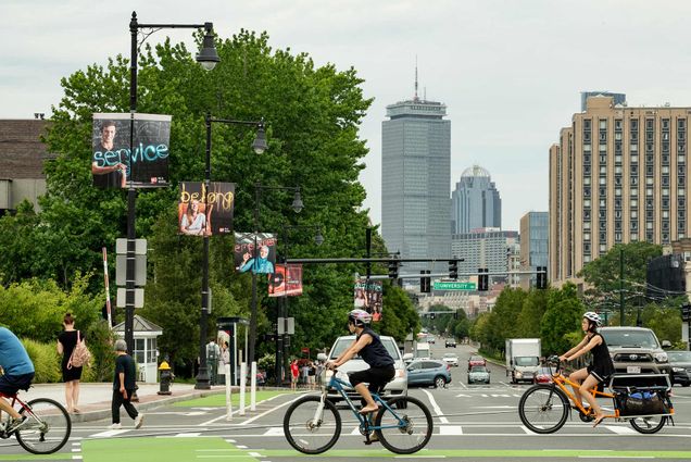 Image of three bikers crossing Comm Ave on a warm summer day. The Prudential Building is seen in the background. BU banners line the street.