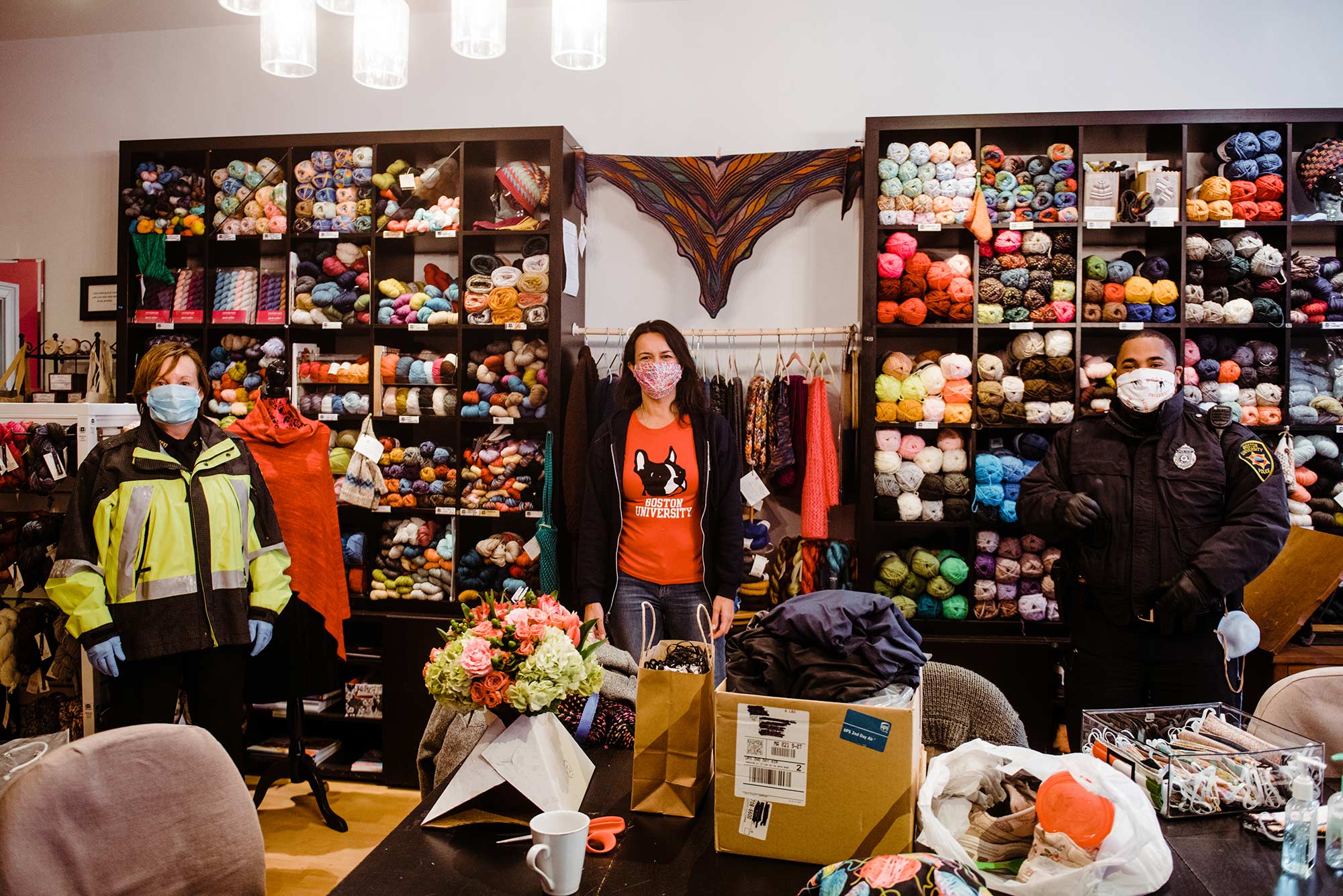Image of Boston City Councilor and BU alum, Annissa Essaibi-George, who has started sew face masks for the Boston University Police Department at the Stitch House, a shop in Dorchester which she founded. Two BU PD officers stand at her left and right; the wall behind them is filled with skeins of yarn.