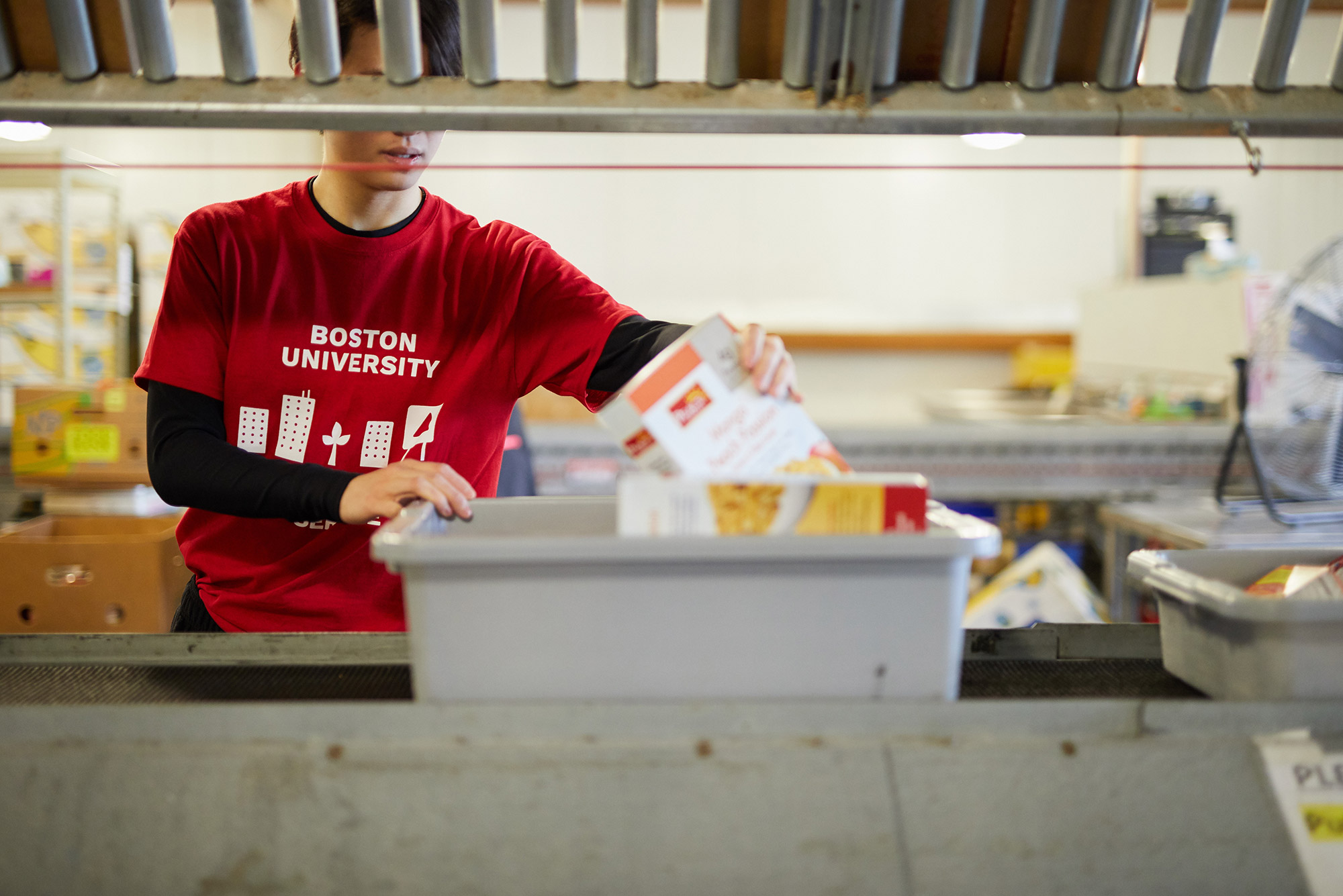 Image of a person wearing a red Boston University shirt volunteering at a food bank; the person's face is hidden by a rack on top of a conveyor belt where they are packing food.