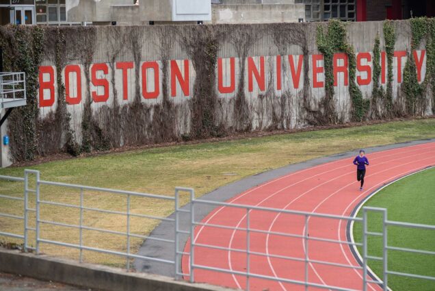 Shot of Nickerson Field with a lone runner in purple jacket running on the track in March of 2020. "Boston University" is seen written on a wall on one side of the track.
