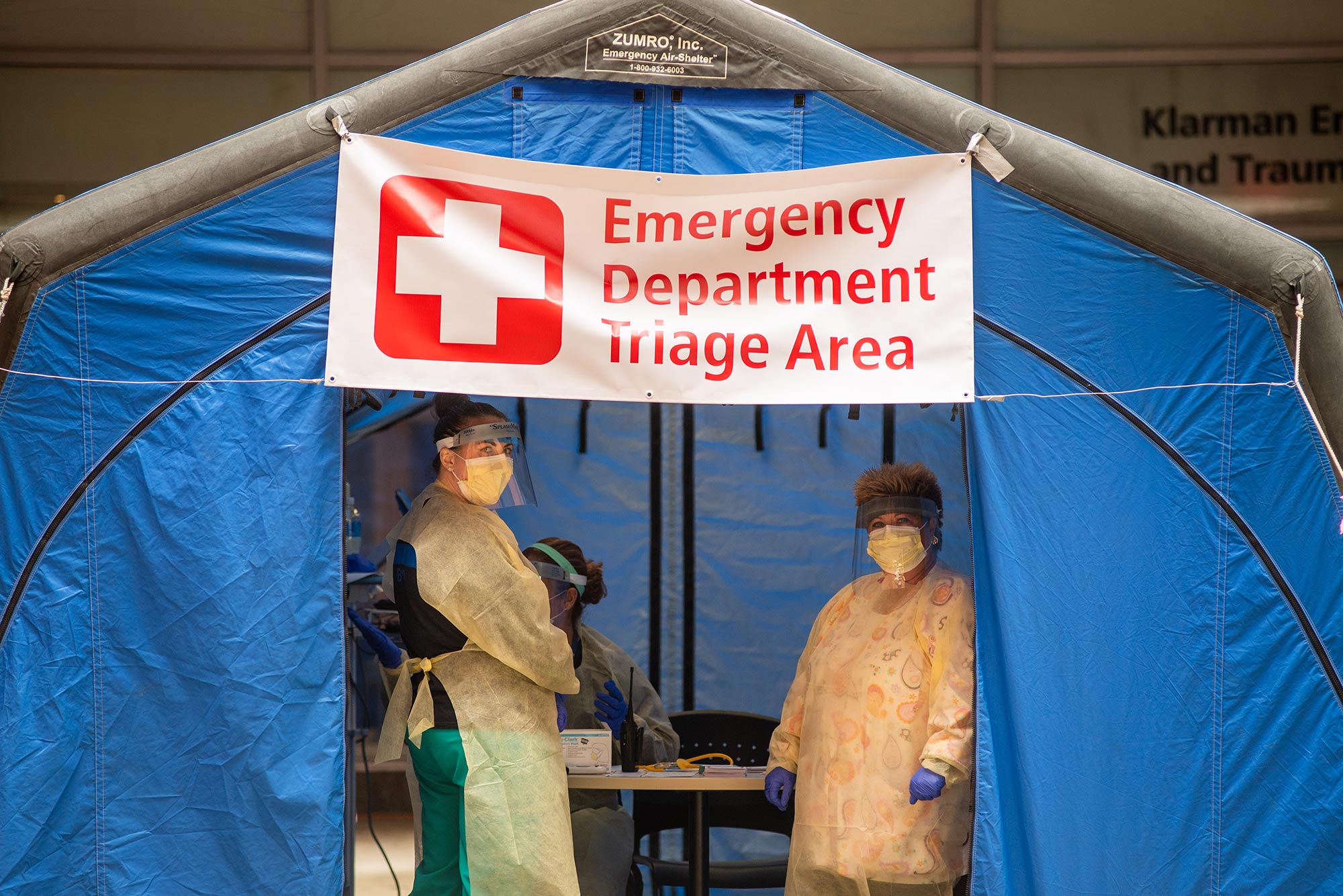 Image of Marisa McIntyre, RN, left, and Maureen Shanahan-Frappier, RN, waiting for patients outside Boston Medical Center March 20, 2020 in a blue tent with a sign that says "Emergency Department Triage Area."