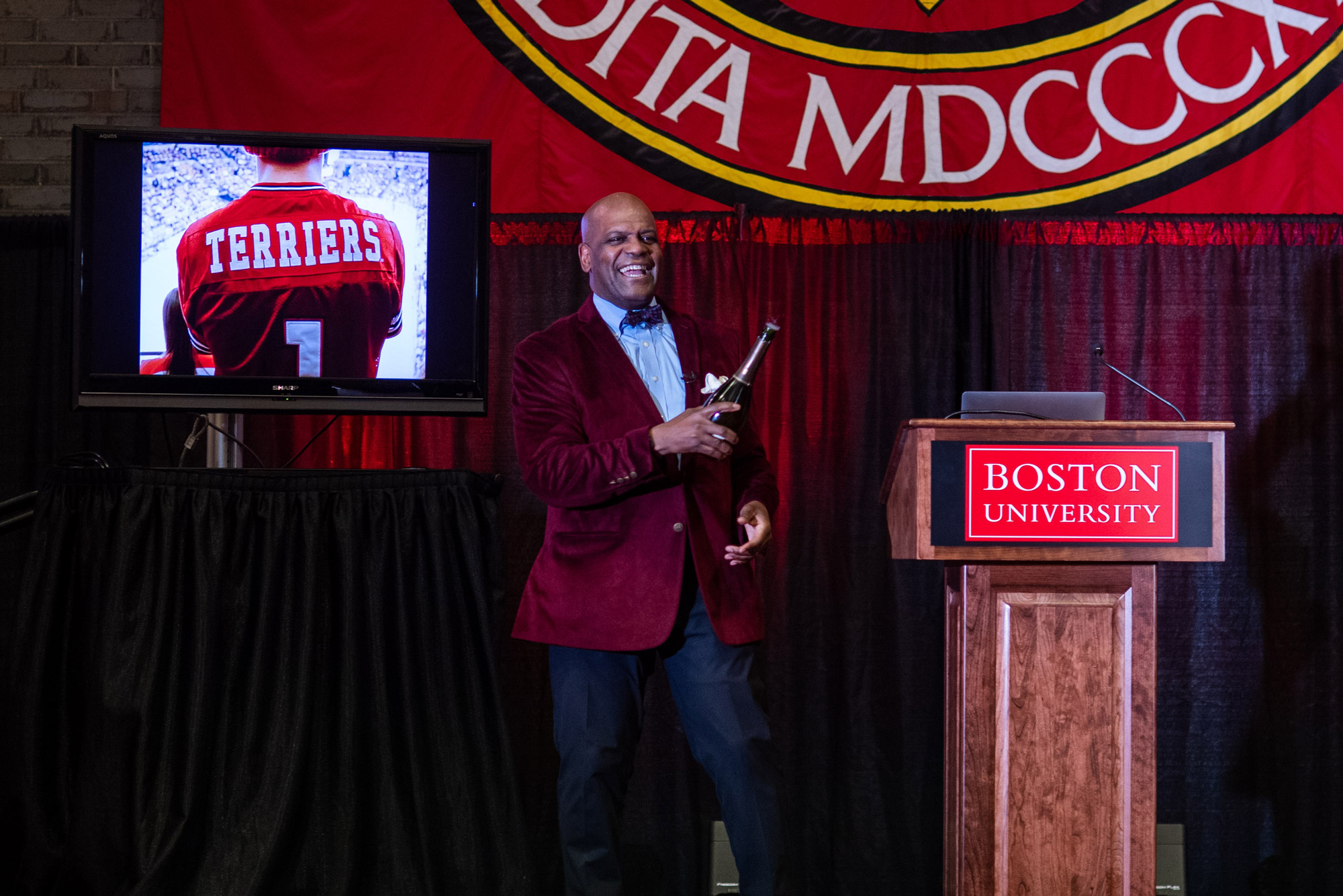 Dean Kenneth Elmore stands on a stage in the George Sherman Union next to a podium with a red Boston University sign on it. He wears a red blazer and holds a champagne bottle. Behind him, a screen shows an image of the back of a Terriers jersey.