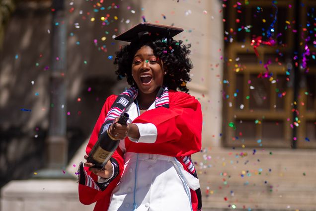 Ann-Lyssa Asare (Pardee’20) pops a champagne bottle full of confetti for a photo shoot in Marsh Plaza May 13, 2020. She wears a graduation cap and a red robe.