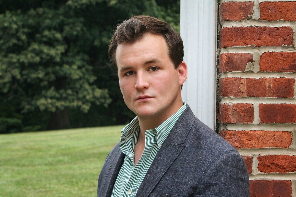 Portrait of Eric Carey in a blazer and button down shirt. Behind him is a brick wall and grass. 