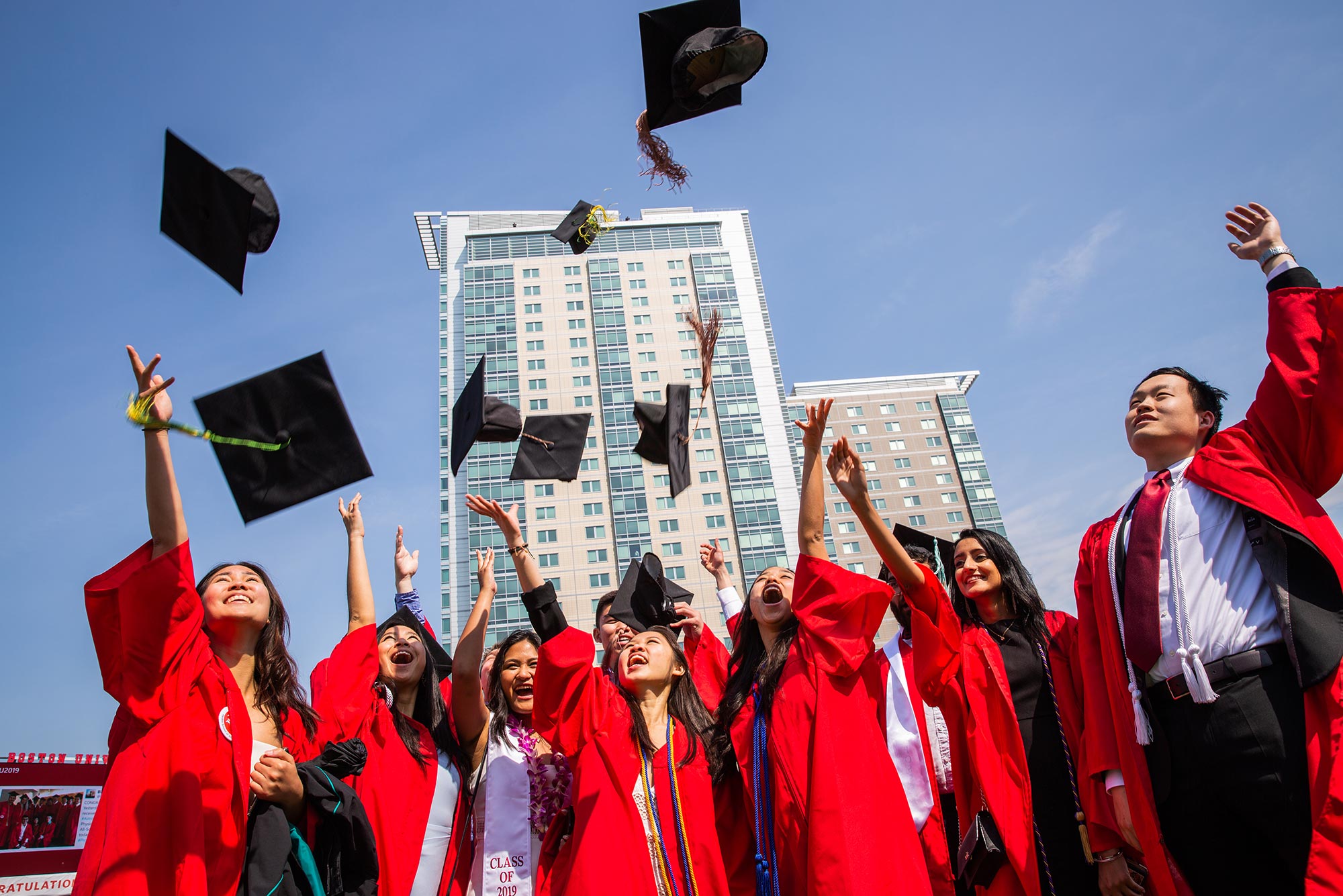 Photo of 2019 graduates at Boston University's Commencement on Nickerson Field on May 19, 2019 throwing their hats into the air. They are wearing red gowns. It's a sunny day and the sky is blue.