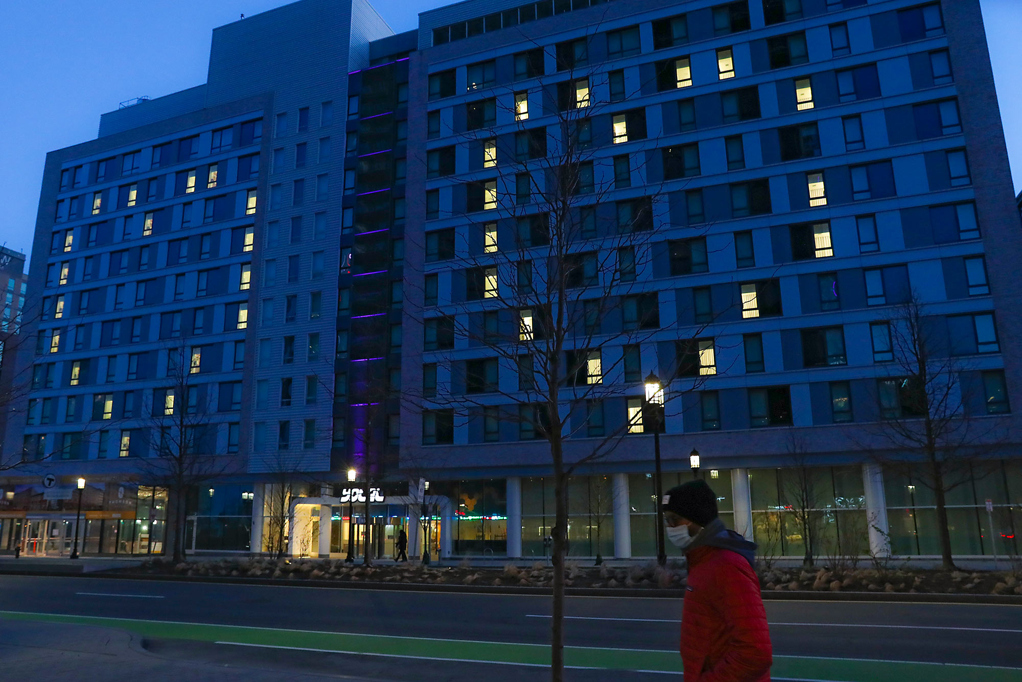 Photo of a lone pedestrian in a red sweater and a mask, walking past the Yotel building on Seaport Boulevard in Boston at sundown on April 22, 2020. Since the start of the coronavirus outbreak, the hotel has the rooms light up in the shape of a heart; two hearts are seen in the background.