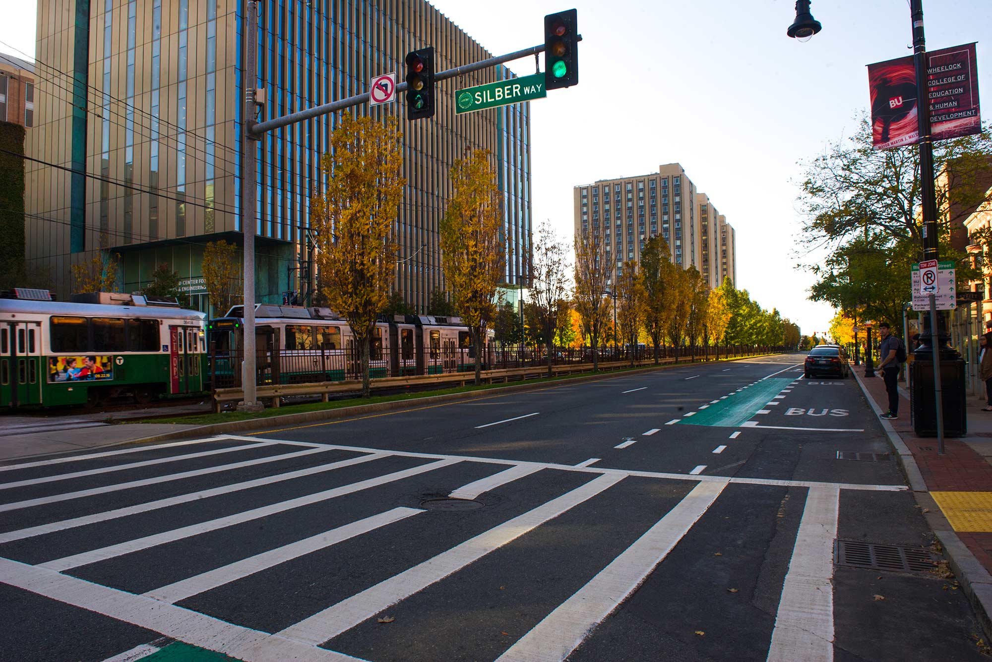 Photo of Commonwealth Avenue in East Campus by Silber Way. The T passes by and the trees look golden. Students wait at the BU BUS stop. Warren towers and Wheelock College banners are seen in the background.