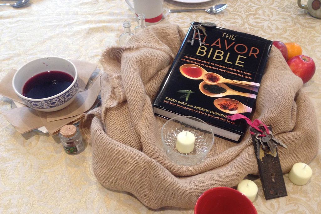 Image of a book, bowl of wine, candles, and burlap sack on a table. The book is titled "The Flavor Bible." This image was taken during a student-prepared meal from a class taught by Metcalf award winner Courtney Goto.