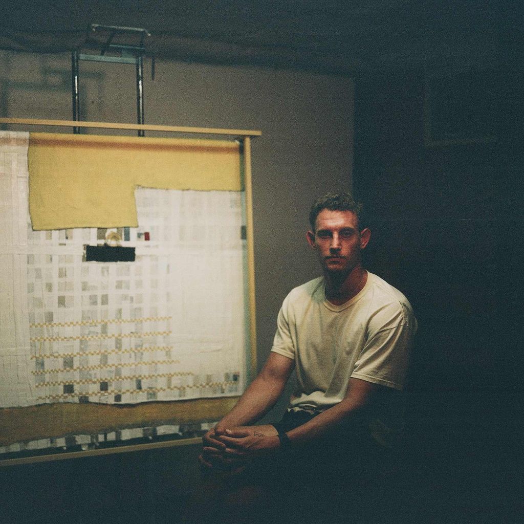 Photo of Alex Stern in his studio next to an easel. He sits with his hands folded across his knee.