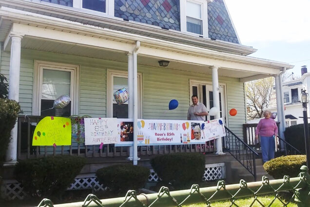 Image of Rose's front porch, she stands on the steps and a man stands near the door. The BU Dining Services team has hung a sign that says "Happy Birthday, Rose's 85th birthday!" and tied balloons to her porch. Photo courtesy of Michael Jackson
