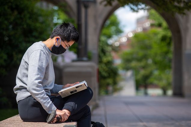 A student wearing a mask sitting and reading a book in Marsh Plaza