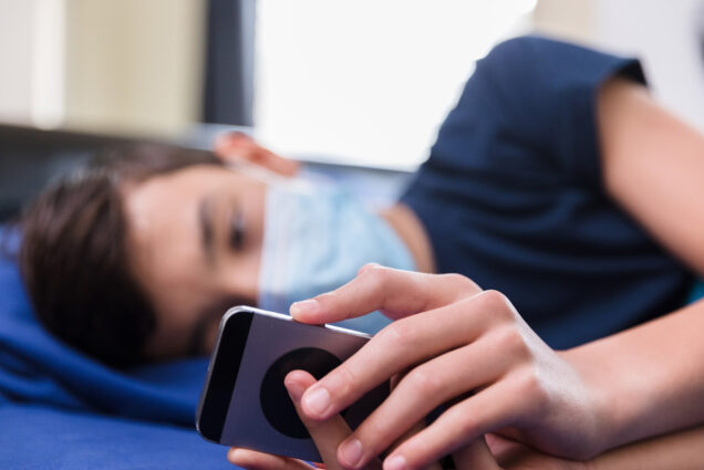 During COVID-19 pandemic, a teen wearing a protective mask lies in bed watching a video on his smart phone. His face is blurred.