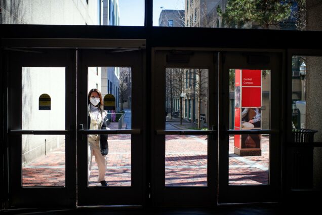 Image of a young person with a white mask opening the side door of the GSU. The hallway and doorframes are dark and it's sunny outside.