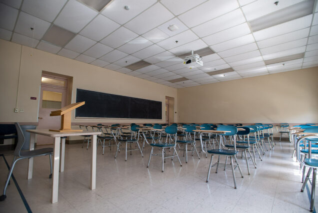 Photo of an empty classroom from inside CAS March 18, 2020, after campus cleared out due to the pandemic. A professor's podium sits at the front of a classroom of rows of teal single-person desks.