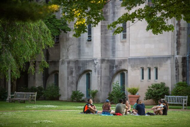 A group of students sit on blankets in the grass behind Marsh Chapel, a tree is seen in the top left of the photo.
