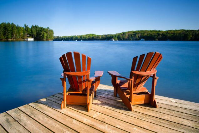 Photo of Adirondack chairs on a wooden dock on a calm lake in Muskoka, Ontario Canada. Cottages nestled between trees are visible across the water.